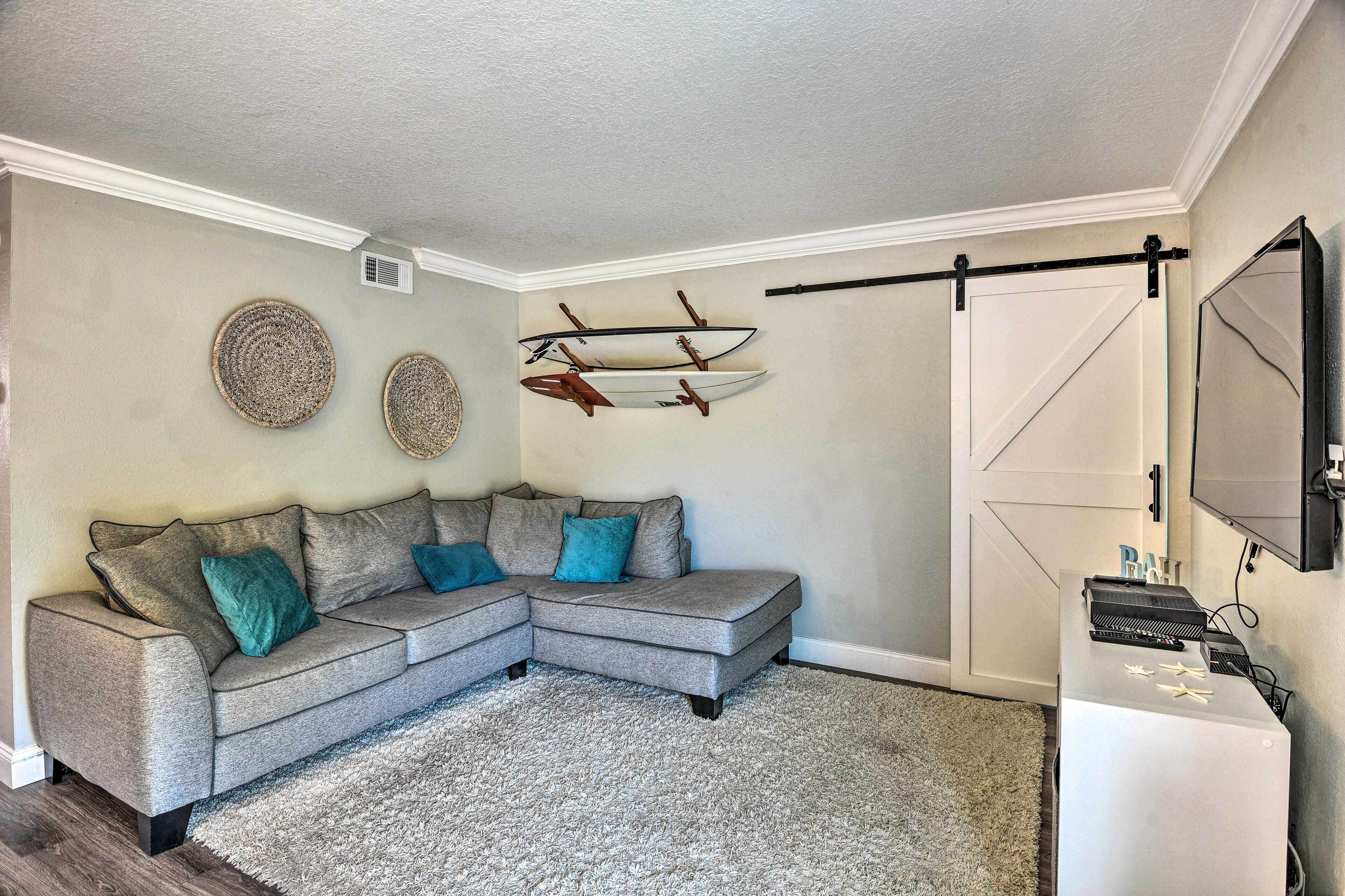 A living room with a grey sectional sofa, two woven wall decorations, and a mounted surfboard.
