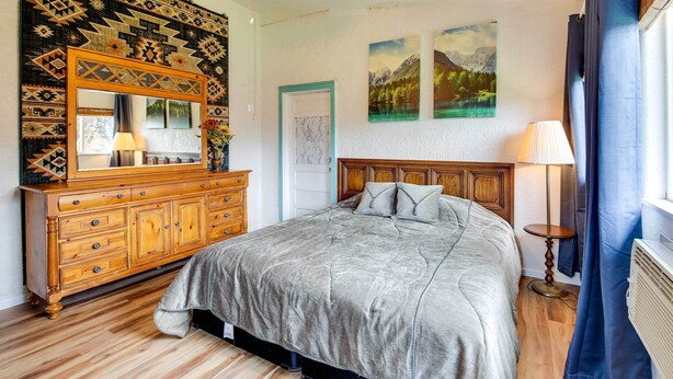 A bedroom with a wooden dresser, a bed with a grey bedspread, a mirror, and two landscape paintings on the wall. - Spacious Wyoming Retreat: Fishing & Hunting (Torrington)
