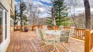 Eine Holzterrasse mit einem Glastisch und Stühlen, umgeben von Bäumen und einem bewölkten Himmel.