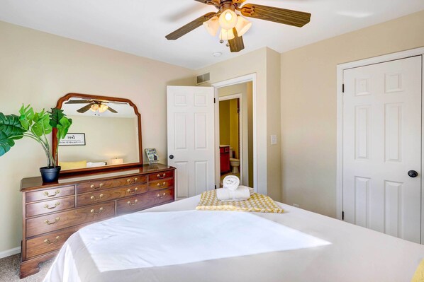 A bedroom with a bed, a dresser, a mirror, a ceiling fan, and a door leading to a bathroom.