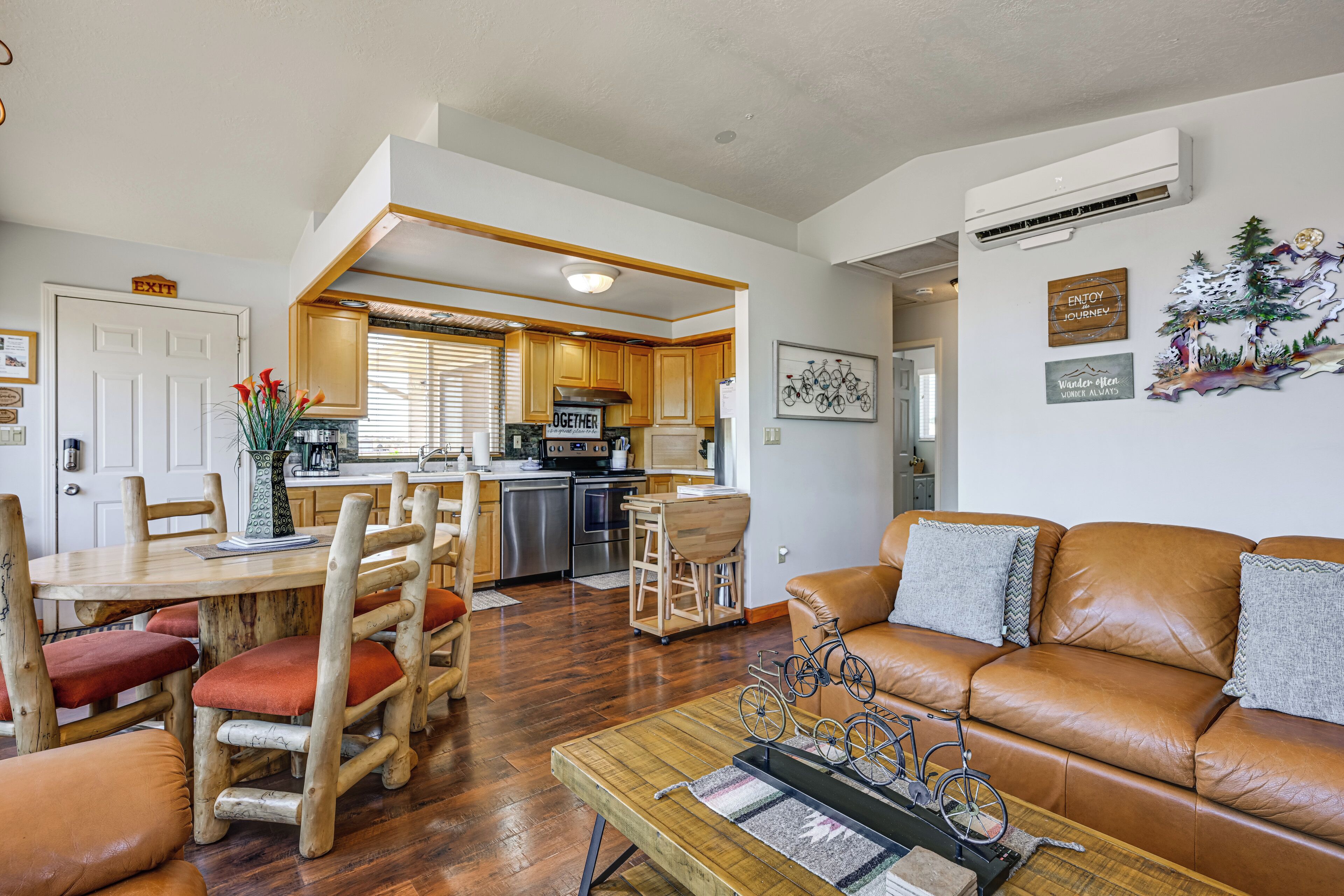 A spacious living area with a leather sofa, wooden dining table, and chairs. A kitchen with wooden cabinets and a refrigerator is visible in the background.