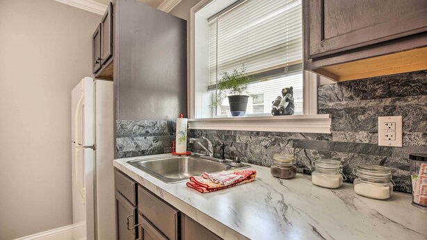 A kitchen with a marble countertop, a double sink, dark cabinets, a window with blinds, and a potted plant.