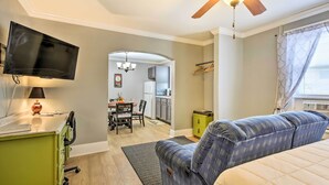 A hotel room with a bed, a desk with a lamp, a TV, a ceiling fan, and a view of the kitchen and dining area.