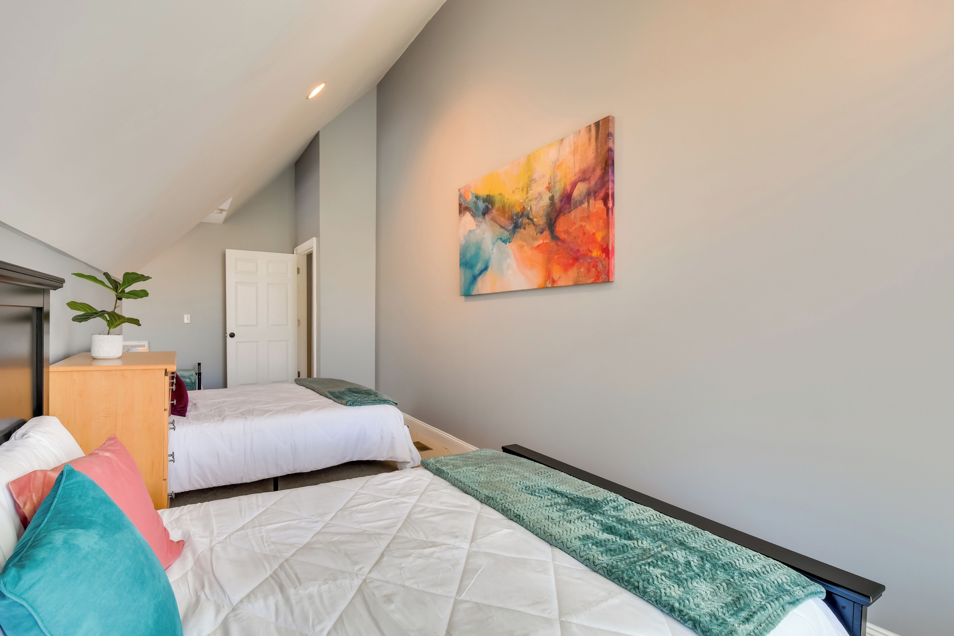 A bedroom with two beds, a wooden nightstand with a plant, a colorful abstract painting on the wall, and a door in the background.