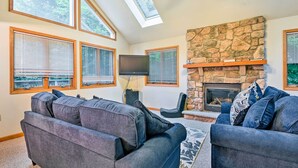 A living room with a stone fireplace, large windows, and a flat-screen TV. - Walk to Beach: Beautiful Lake Naomi Retreat! (Pocono Pines)