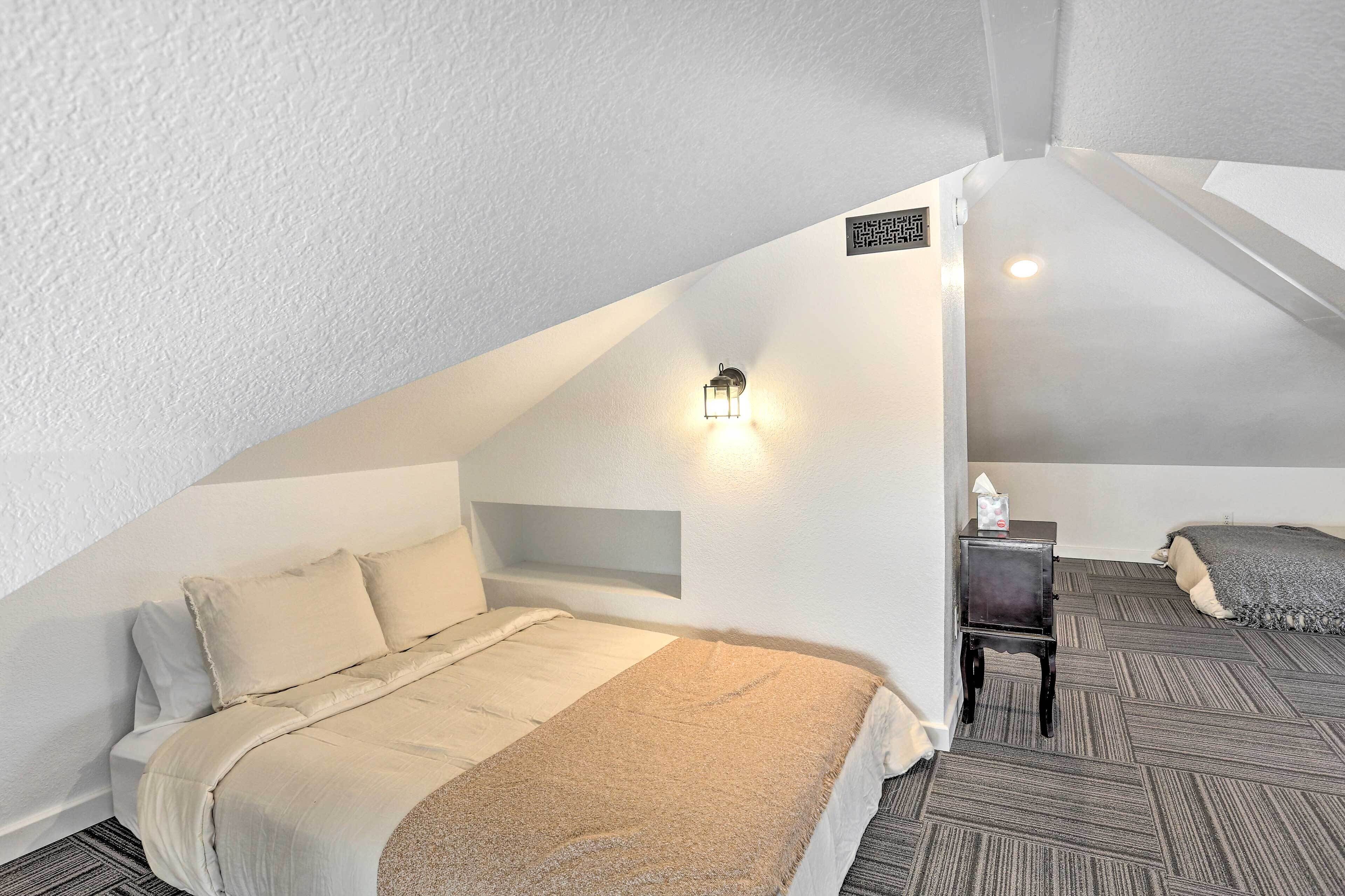 Ett sovrum med ett sluttande tak, en säng med ett beige täcke, ett nattduksbord med en lampa och ett litet bord med en näsdukslåda.