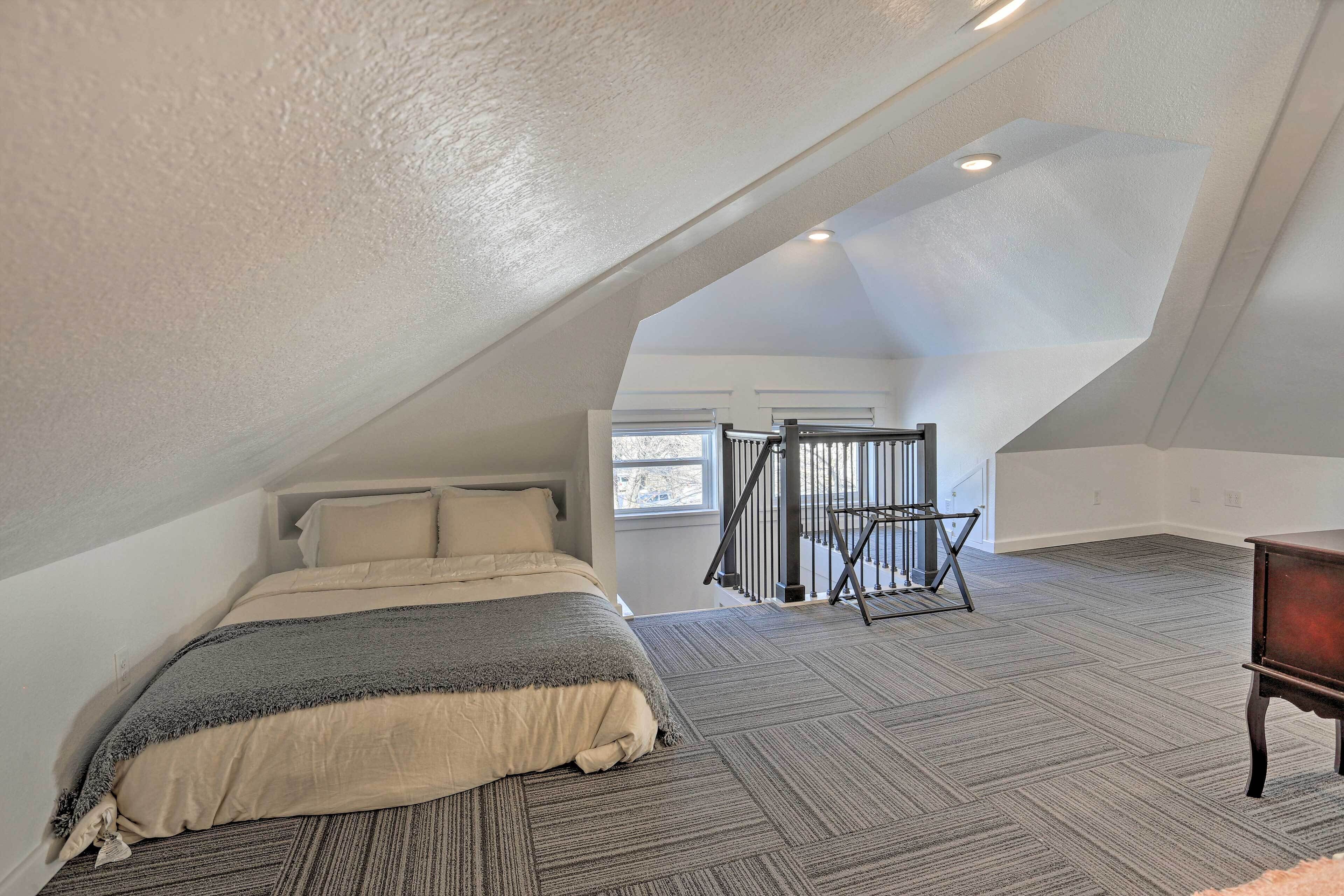 Un dormitorio con techo inclinado, una cama con manta gris y beige, un aparador de madera y un ventanal con vistas al exterior.
