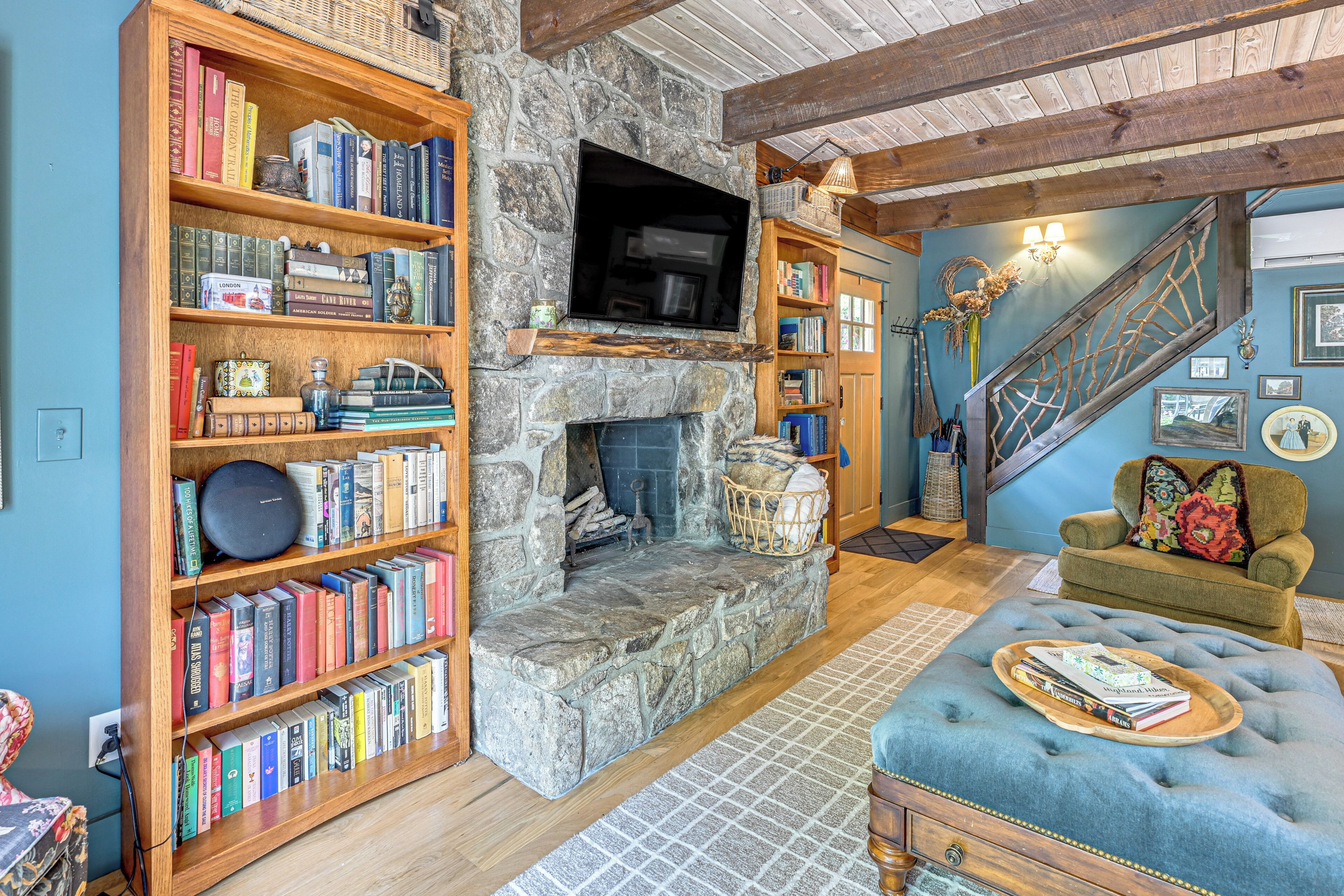 A cozy living room with a stone fireplace, wooden bookshelves, a comfortable blue sofa, and a staircase with a wooden railing.