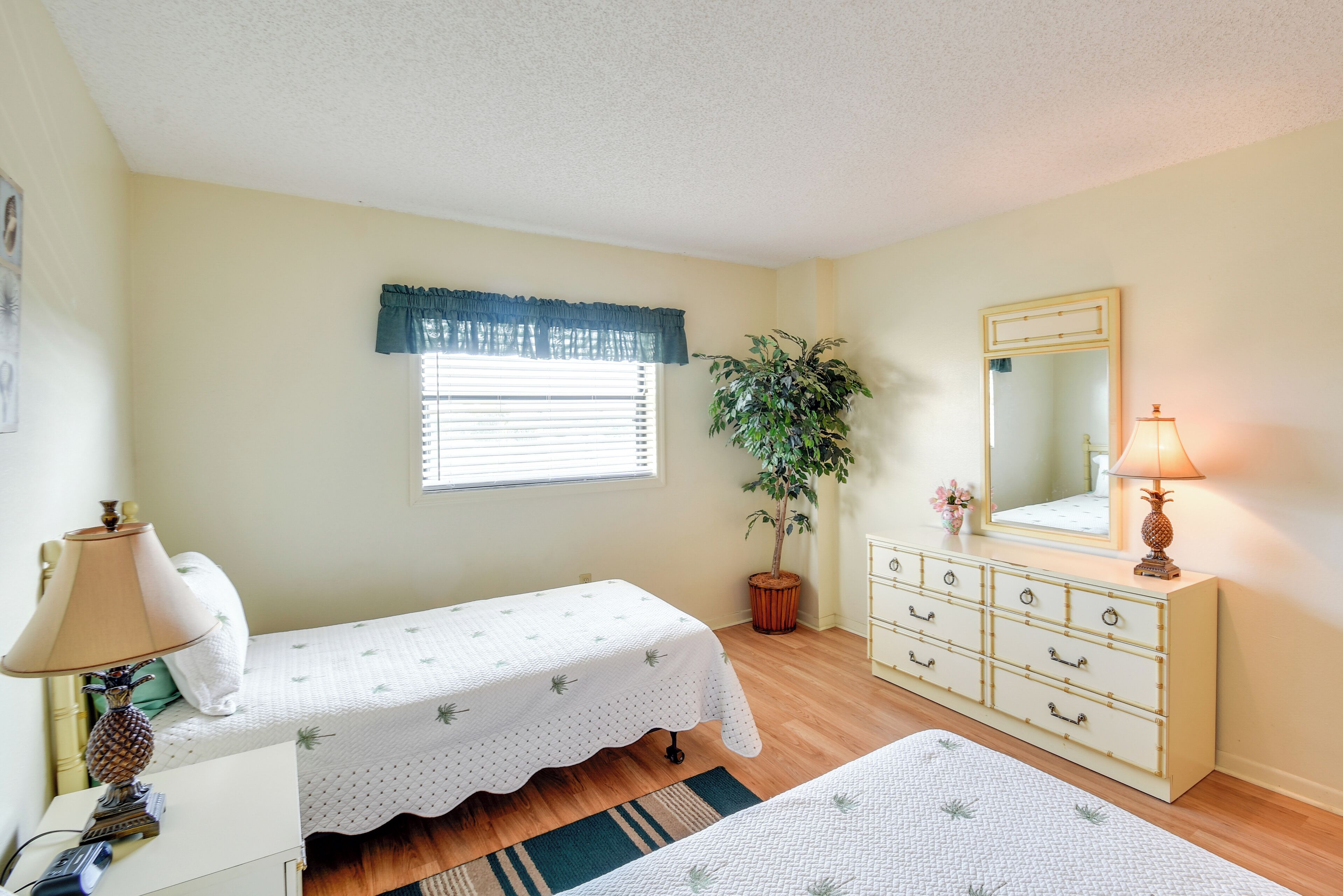 A bedroom with a single bed, a dresser, a mirror, a potted plant, and a window with blinds.