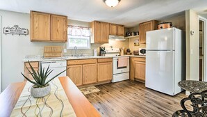 A kitchen with wooden cabinets, a white refrigerator, a stove, and a dining area with a table and chairs. - Cozy House w/ Huge Yard ~ 16 Mi to Fort Bragg (Lillington)