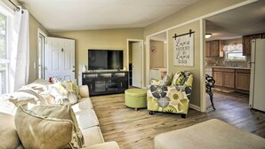 A living room with a sofa, ottoman, and a flat-screen TV. There is a kitchen visible through an open doorway. - Cozy House w/ Huge Yard ~ 16 Mi to Fort Bragg (Lillington)