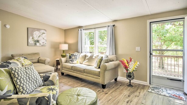 A living room with a beige sofa, a patterned armchair, and a green ottoman. There is a window with curtains, a lamp, and a vase with flowers. - Cozy House w/ Huge Yard ~ 16 Mi to Fort Bragg (Lillington)