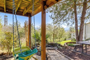 Un porche cubierto con una hamaca, una mesa de picnic y vistas a un jardín y montañas.