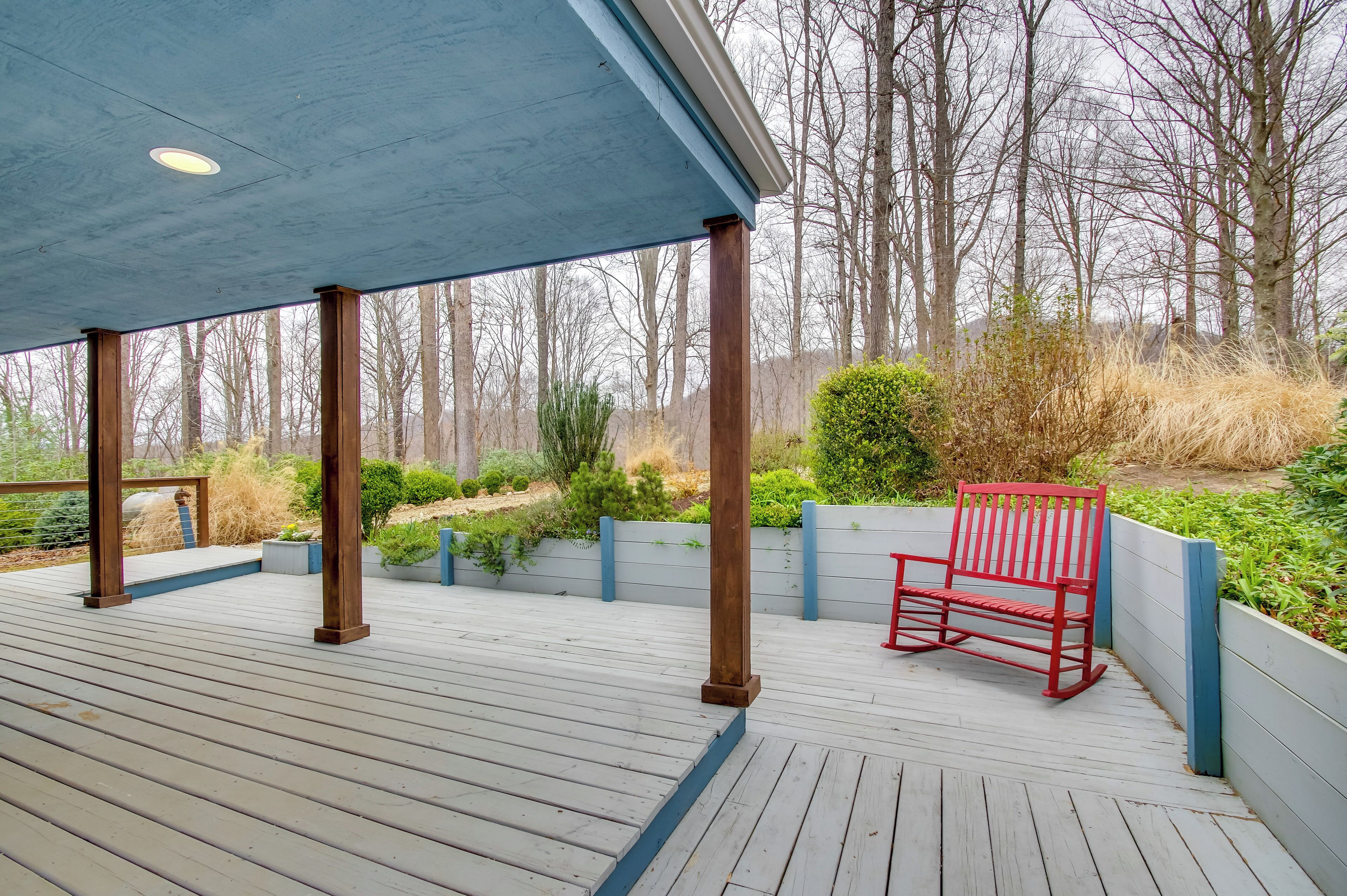 A covered wooden deck with a red bench, surrounded by a garden with various plants and trees.
