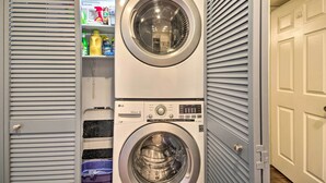 Un cuarto de lavandería con dos lavadoras apiladas de carga frontal, un estante con productos de limpieza y una puerta con picaporte.