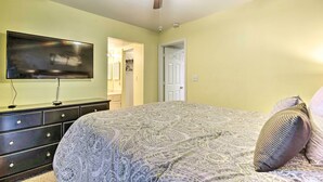 Un dormitorio con una cama, ventilador de techo, televisor sobre un aparador y puerta que da a otra habitación.