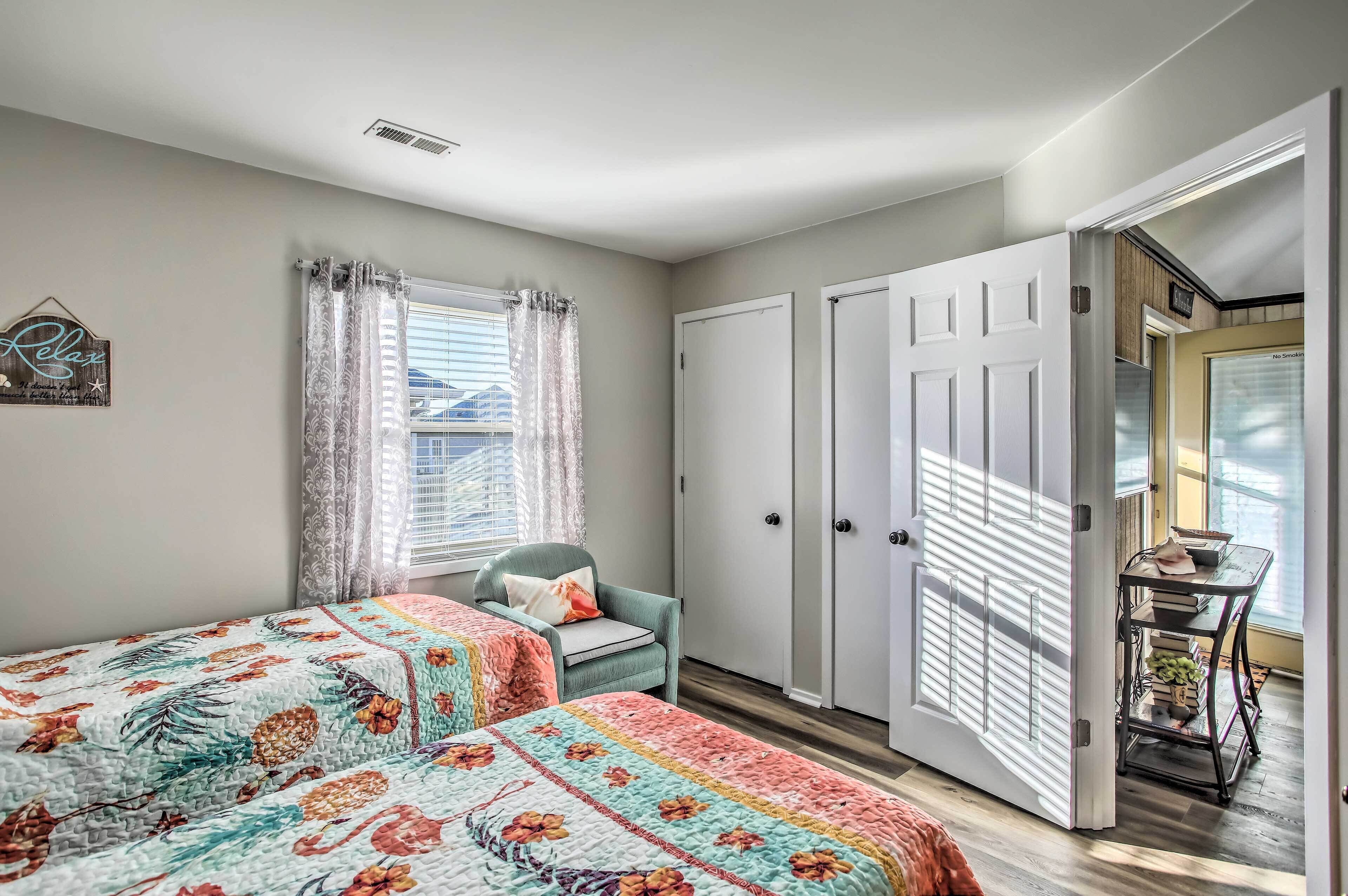 A bedroom with two beds, a chair, a desk with books, and a door leading to another room.
