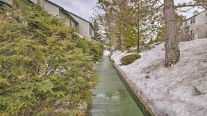 A narrow pathway flanked by snow-covered ground and dense evergreen trees, leading to a building with a sloped roof and a visible staircase. - Lake Placid Home ~ 9 Mi to Whiteface Mtn! (Lake Placid)