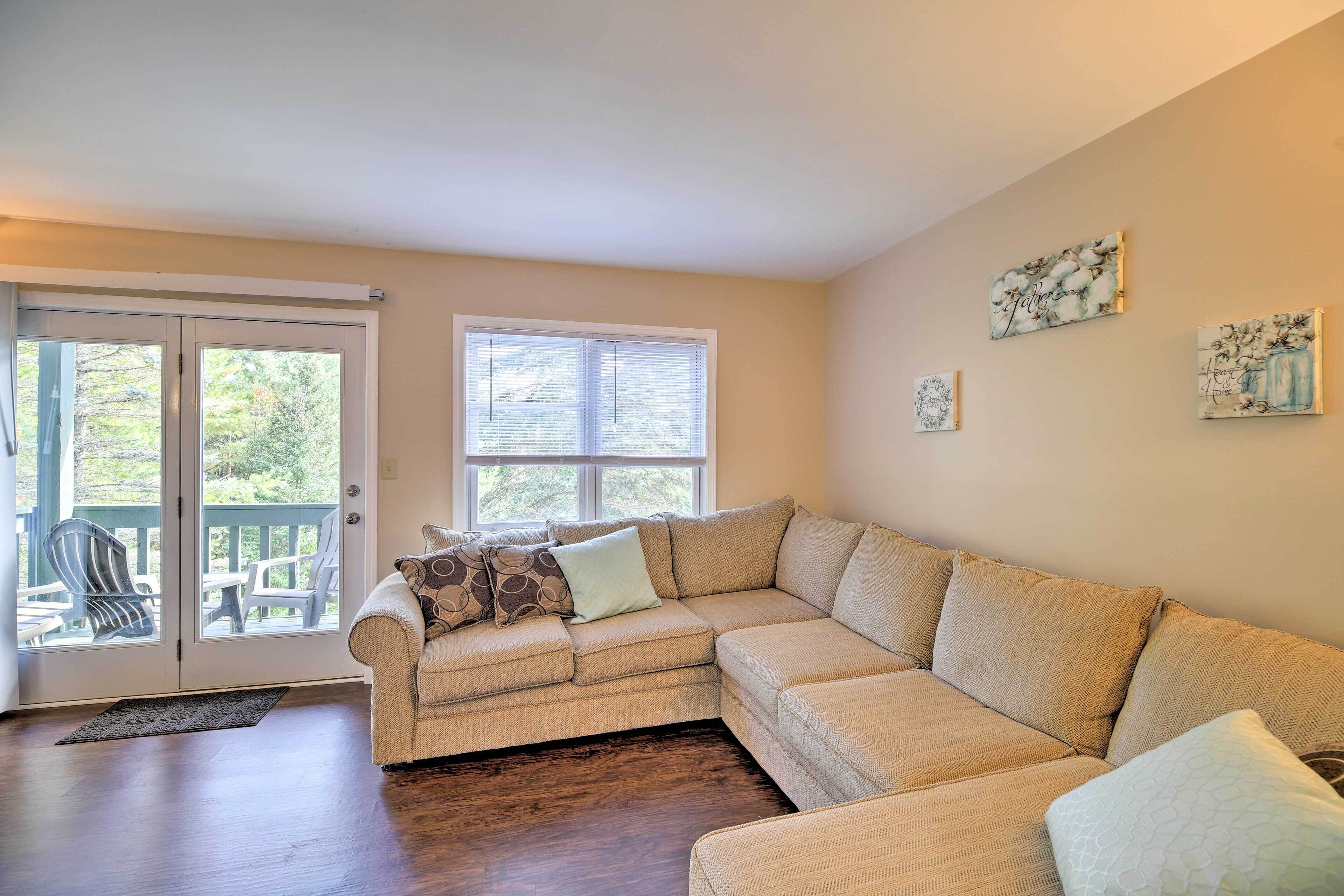 A living room with a beige sectional sofa, a glass door leading to an outdoor deck, and two wall-mounted artworks.
