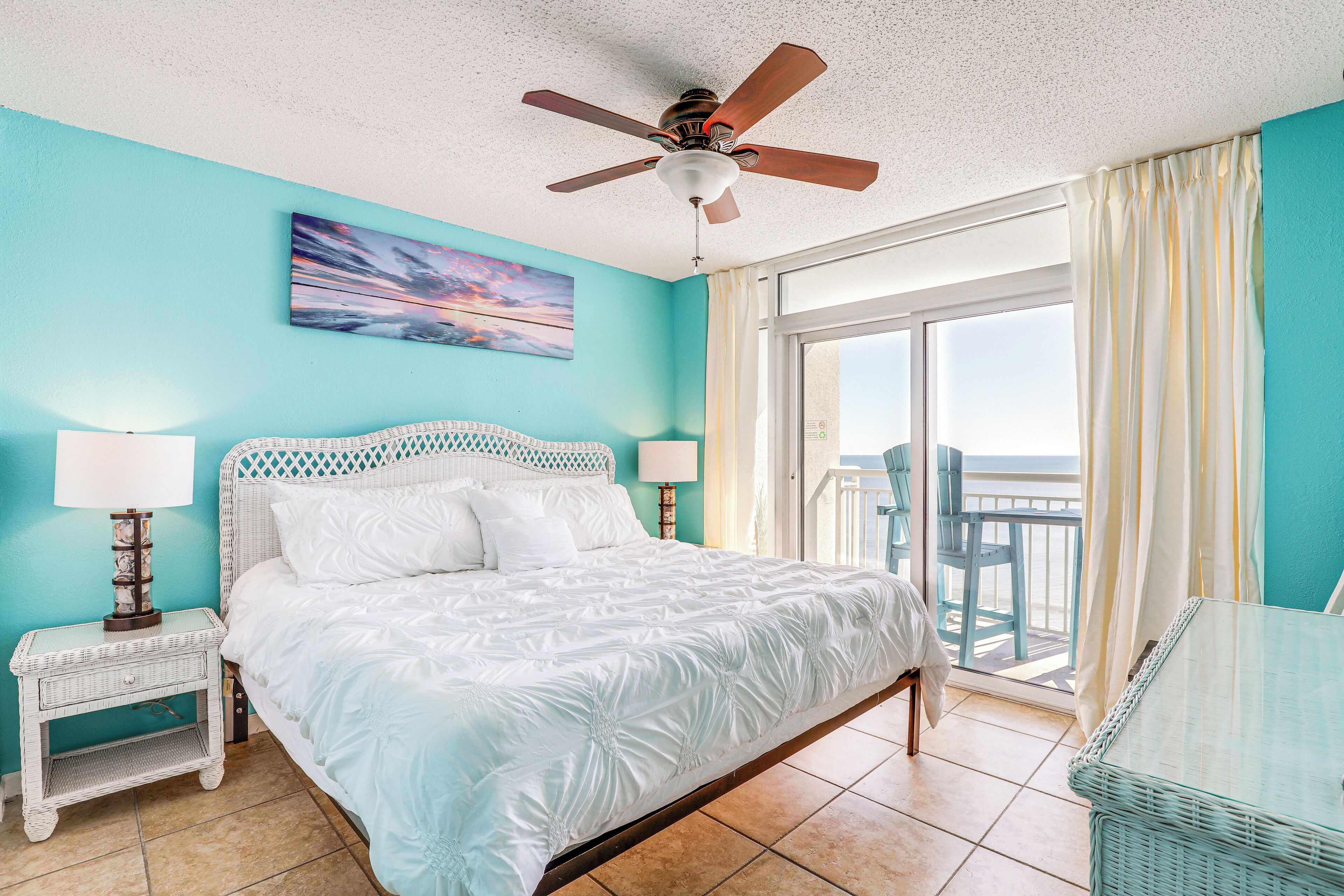 A bedroom with a bed, a ceiling fan, a nightstand, a table, and a view of the outside.