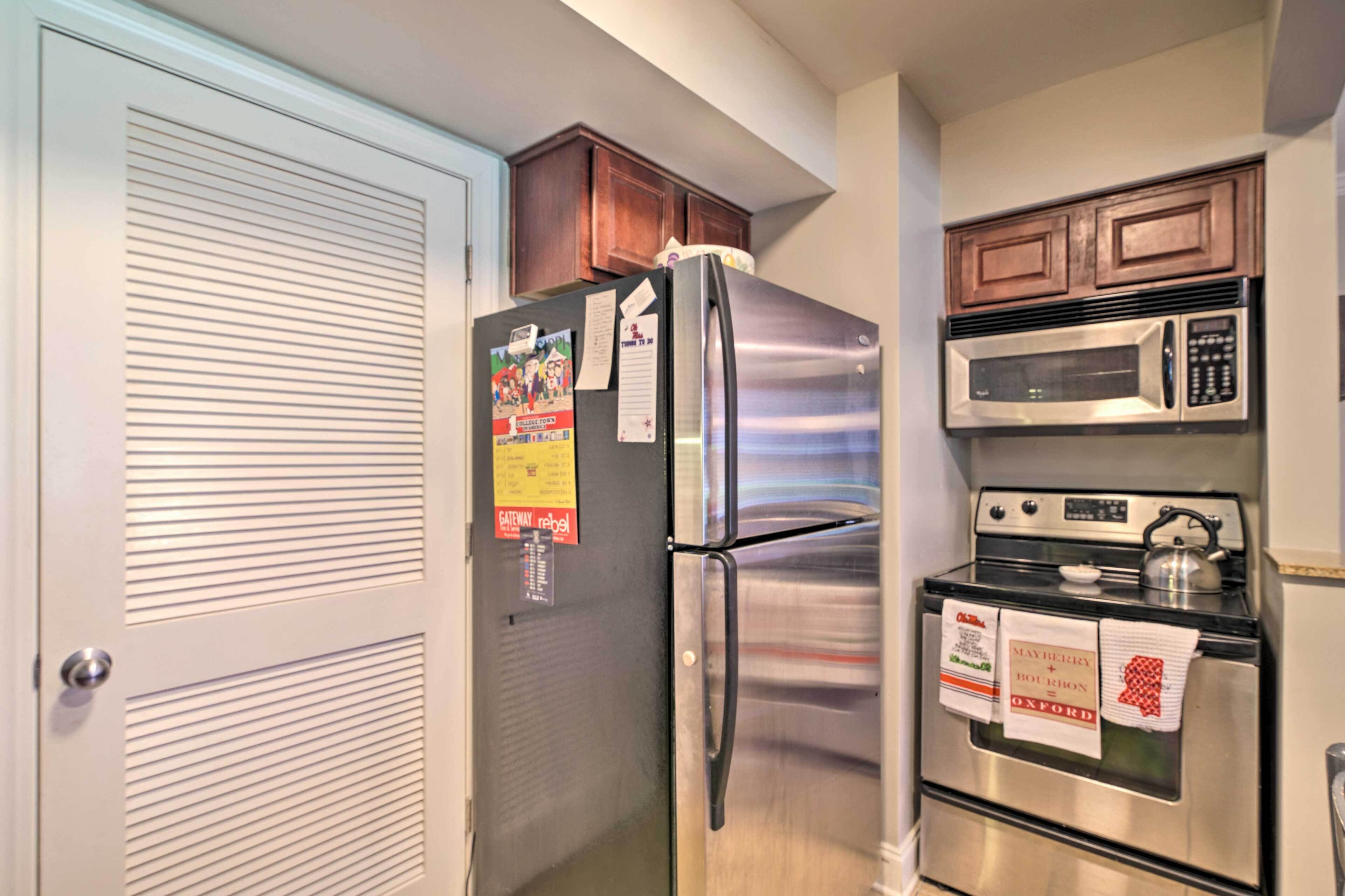 Cocina compacta con un refrigerador de acero inoxidable, un microondas y un horno. Hay armarios de madera sobre los electrodomésticos y una puerta con diseño persiana.