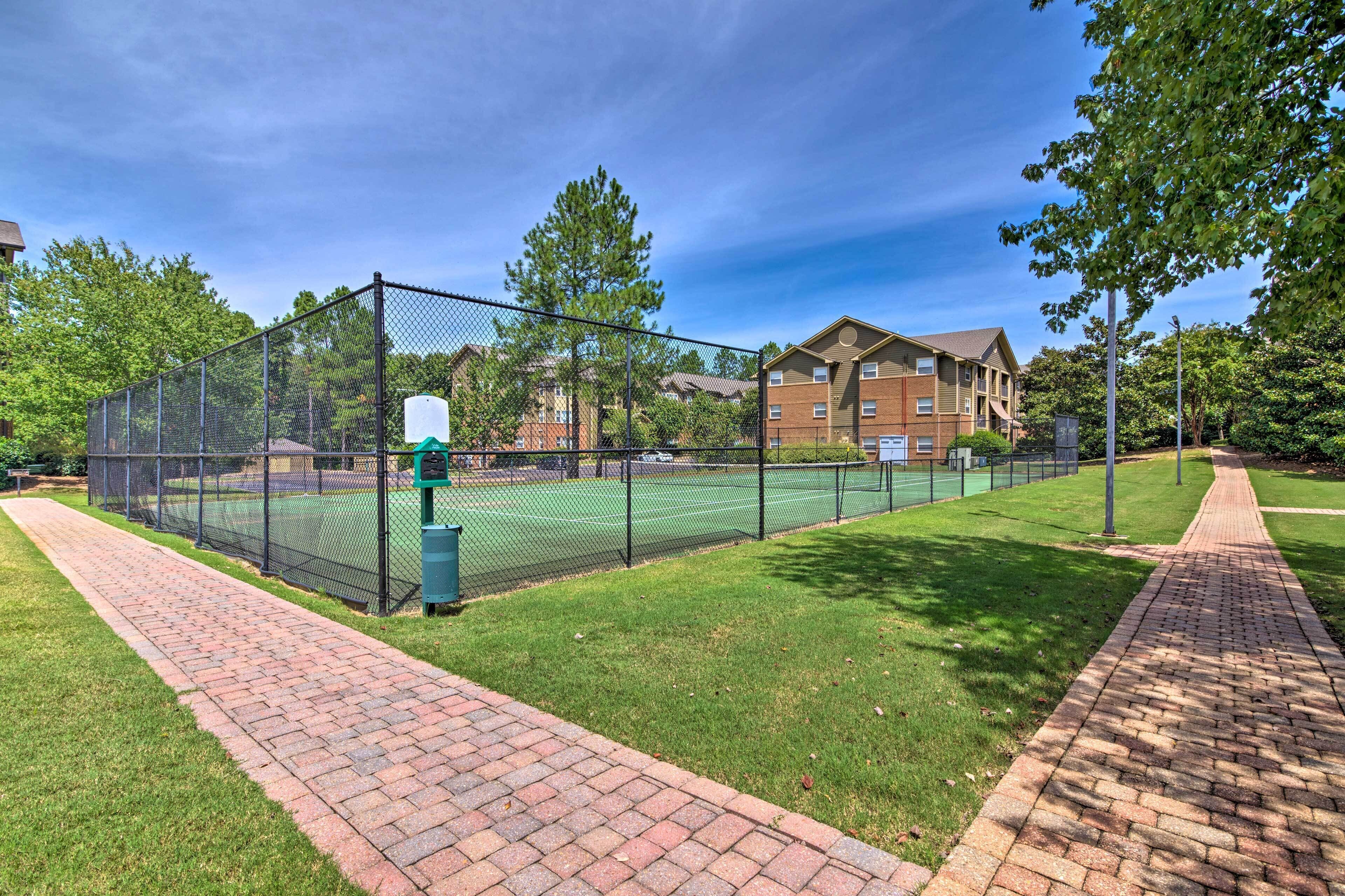Una cancha de tenis rodeada por una cerca de malla metálica, un camino de ladrillo y edificios residenciales al fondo.