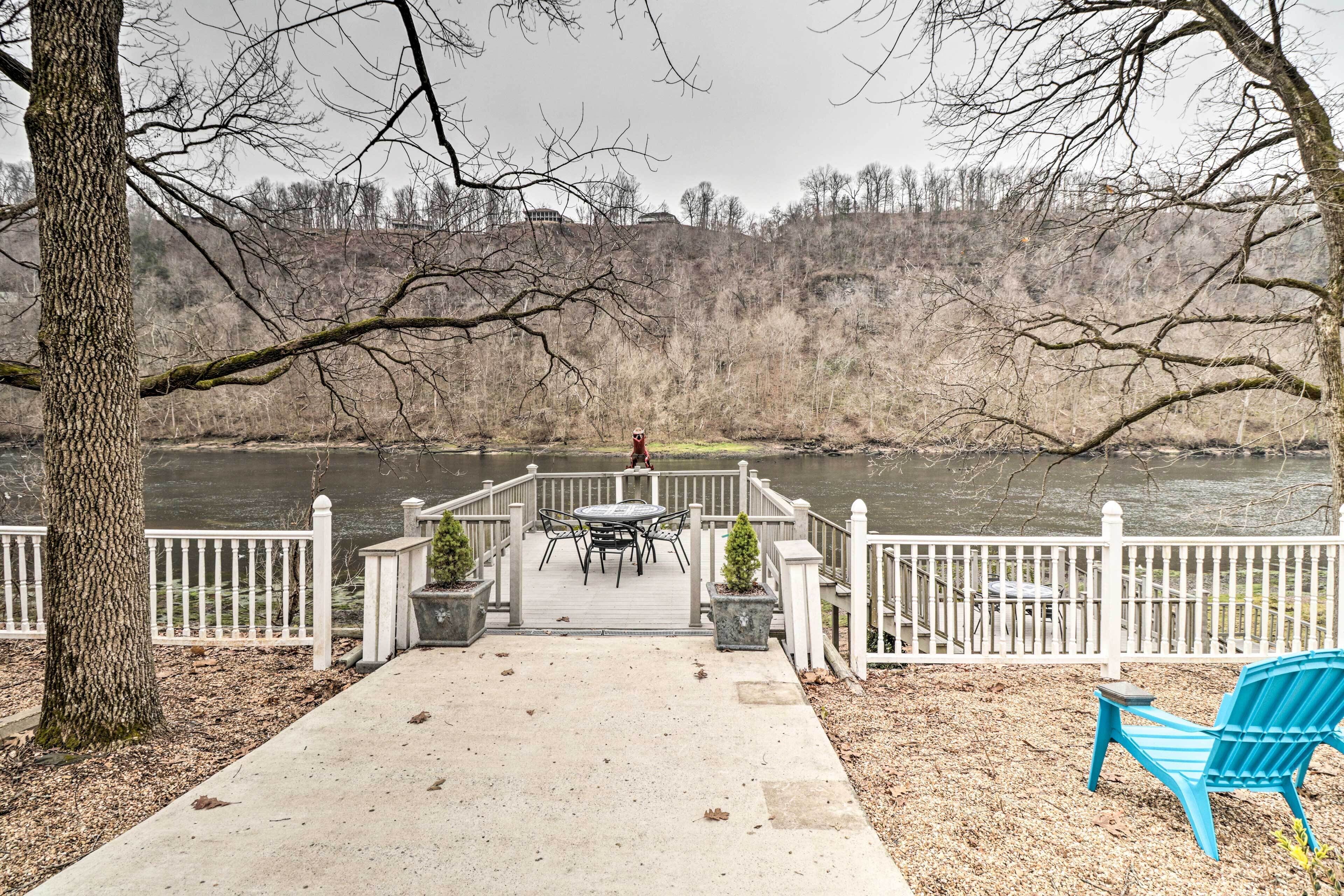 A deck with a table and chairs overlooking a river, surrounded by a white fence and trees.