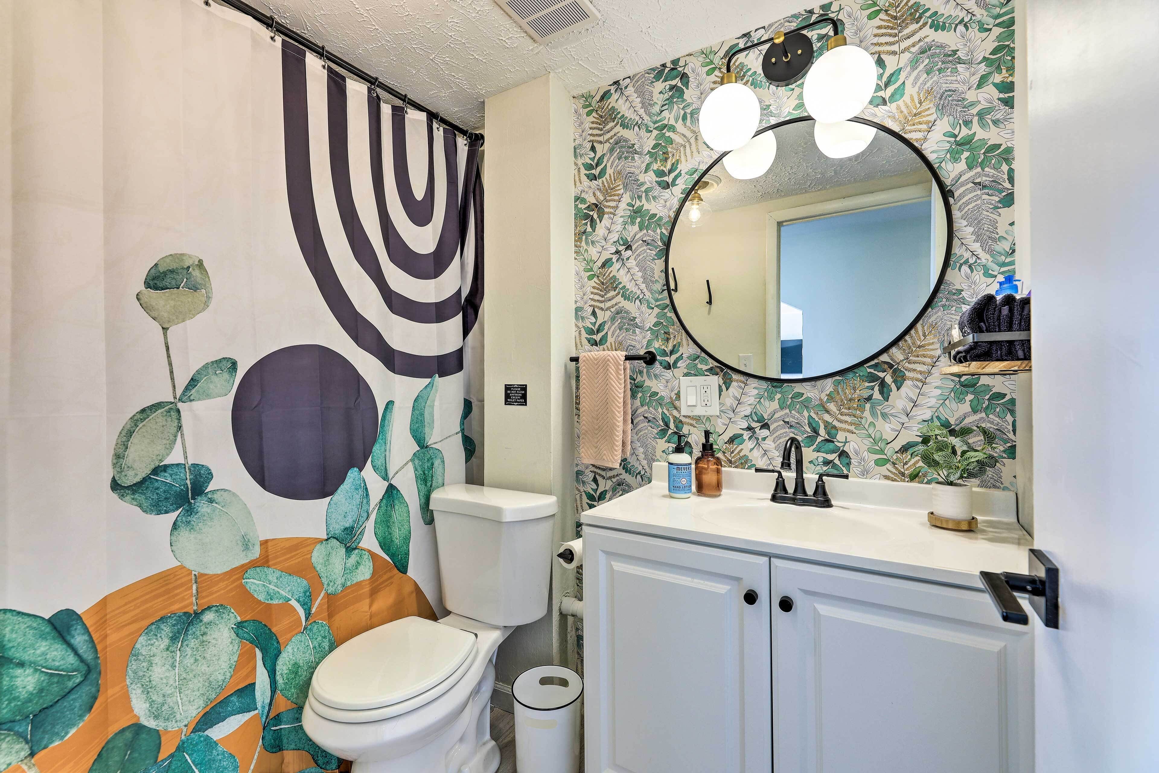 Une salle de bain comprenant des toilettes, un lavabo avec un miroir et un rideau de douche orné d’un motif de feuilles.