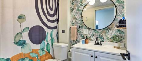 Une salle de bain comprenant des toilettes, un lavabo avec un miroir et un rideau de douche orné d’un motif de feuilles.