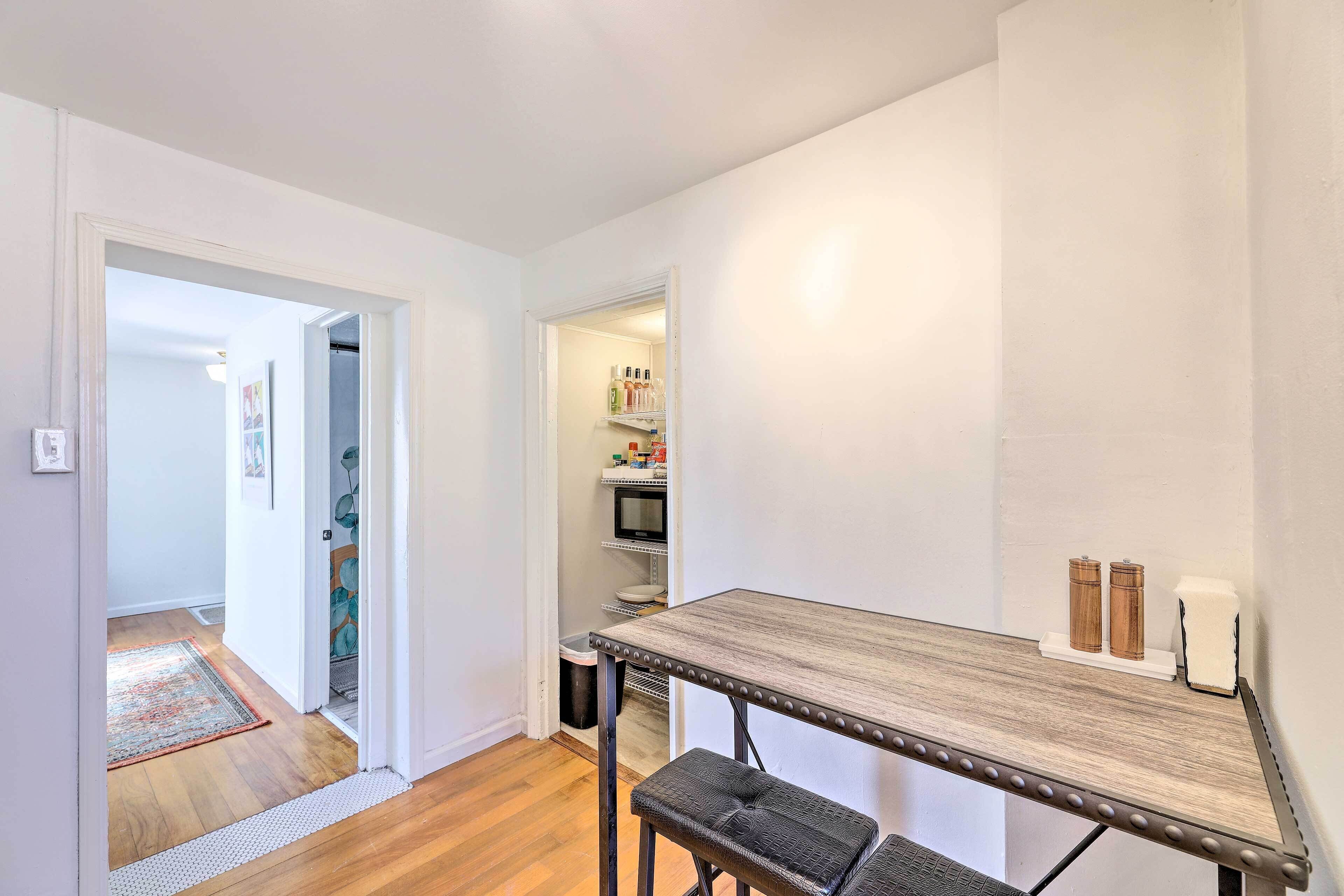 Une petite cuisine avec une table en bois, un banc et un four à micro-ondes. Une porte donne sur un couloir où se trouve un tapis.