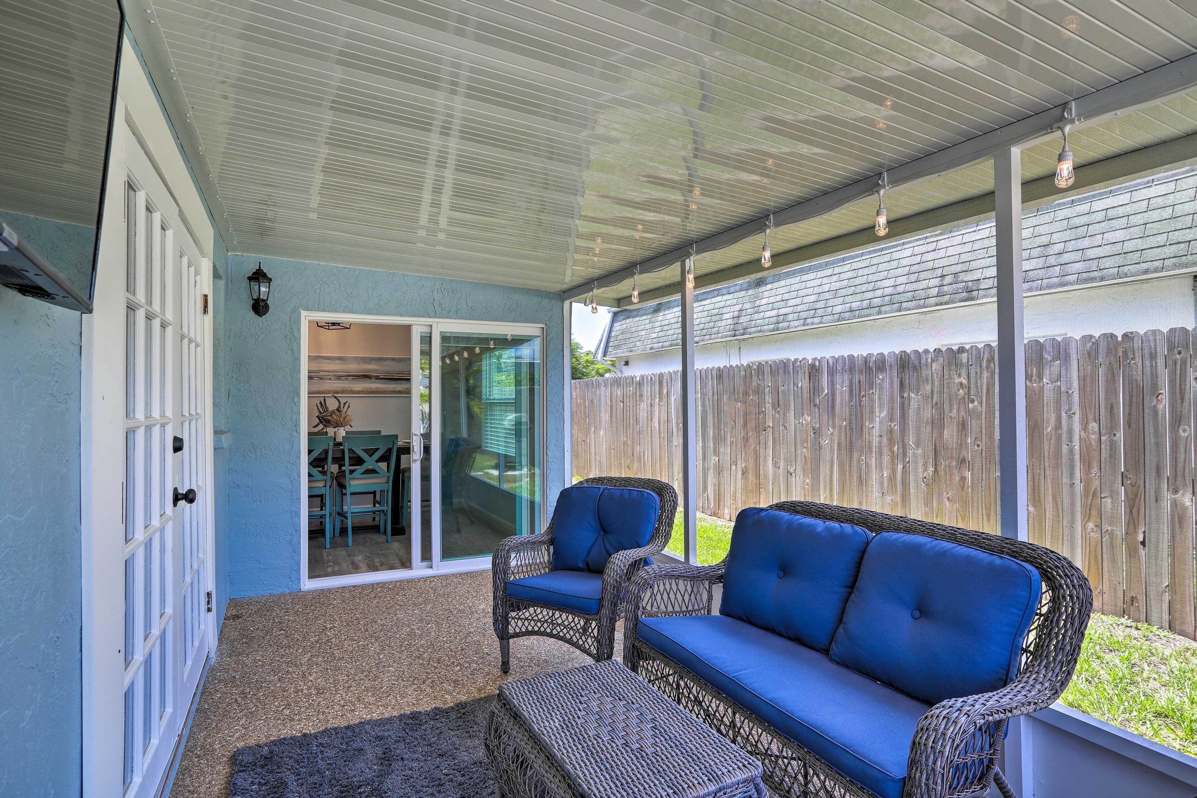 A covered patio area with wicker furniture, a sliding glass door leading to an indoor space, and a wooden fence in the background.