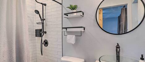 Une salle de bain moderne avec un lavabo en verre, une douche dotée d’un pommeau de douche à pluie et un porte-serviettes agrémenté d’une plante.