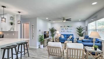 A modern living room with a kitchen area, featuring a blue sofa set, wooden chairs, a dining table, and a bar with stools.