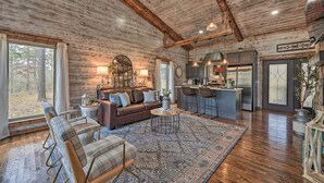 A rustic cabin interior with a wooden ceiling, a cozy living area with a sofa and chairs, a kitchen with bar stools, and large windows overlooking a forest.