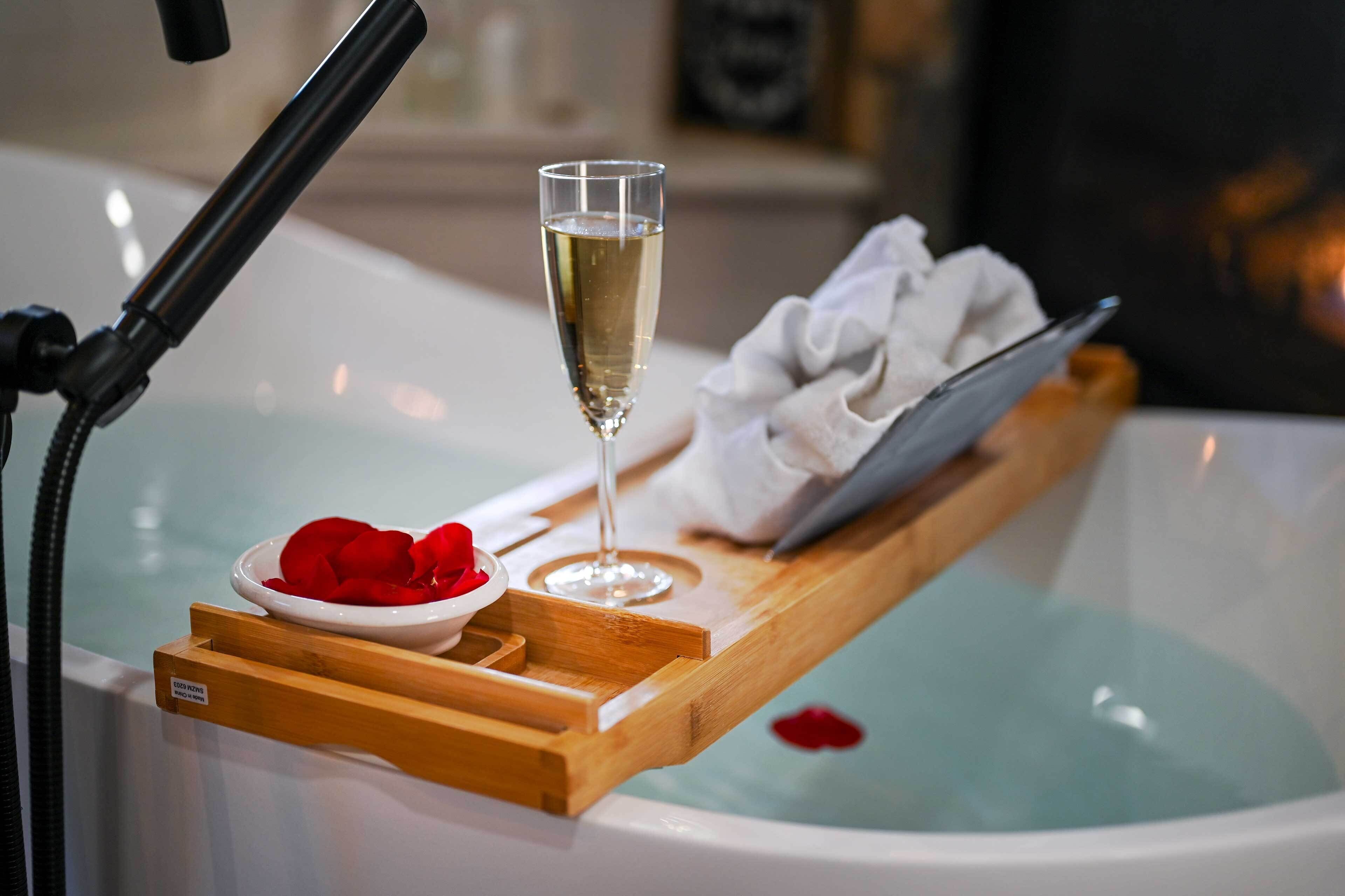 A bathtub with a wooden tray holding a glass of champagne, a bowl of rose petals, and a towel.