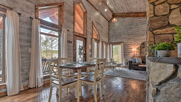 A rustic interior with wooden floors, large windows, a stone wall, and a cozy seating area.