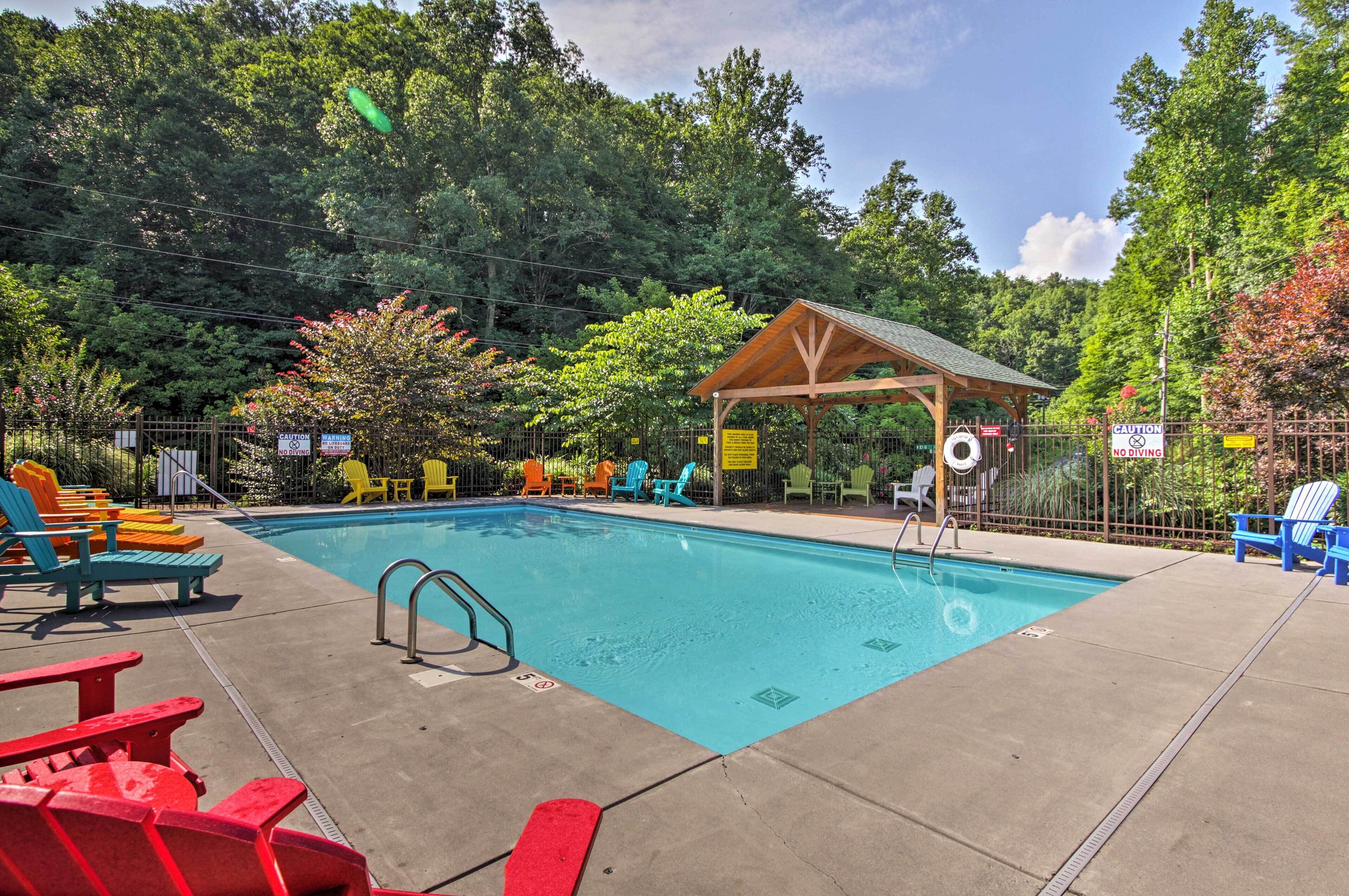 A swimming pool with a wooden pavilion, surrounded by colorful Adirondack chairs and a fenced area.