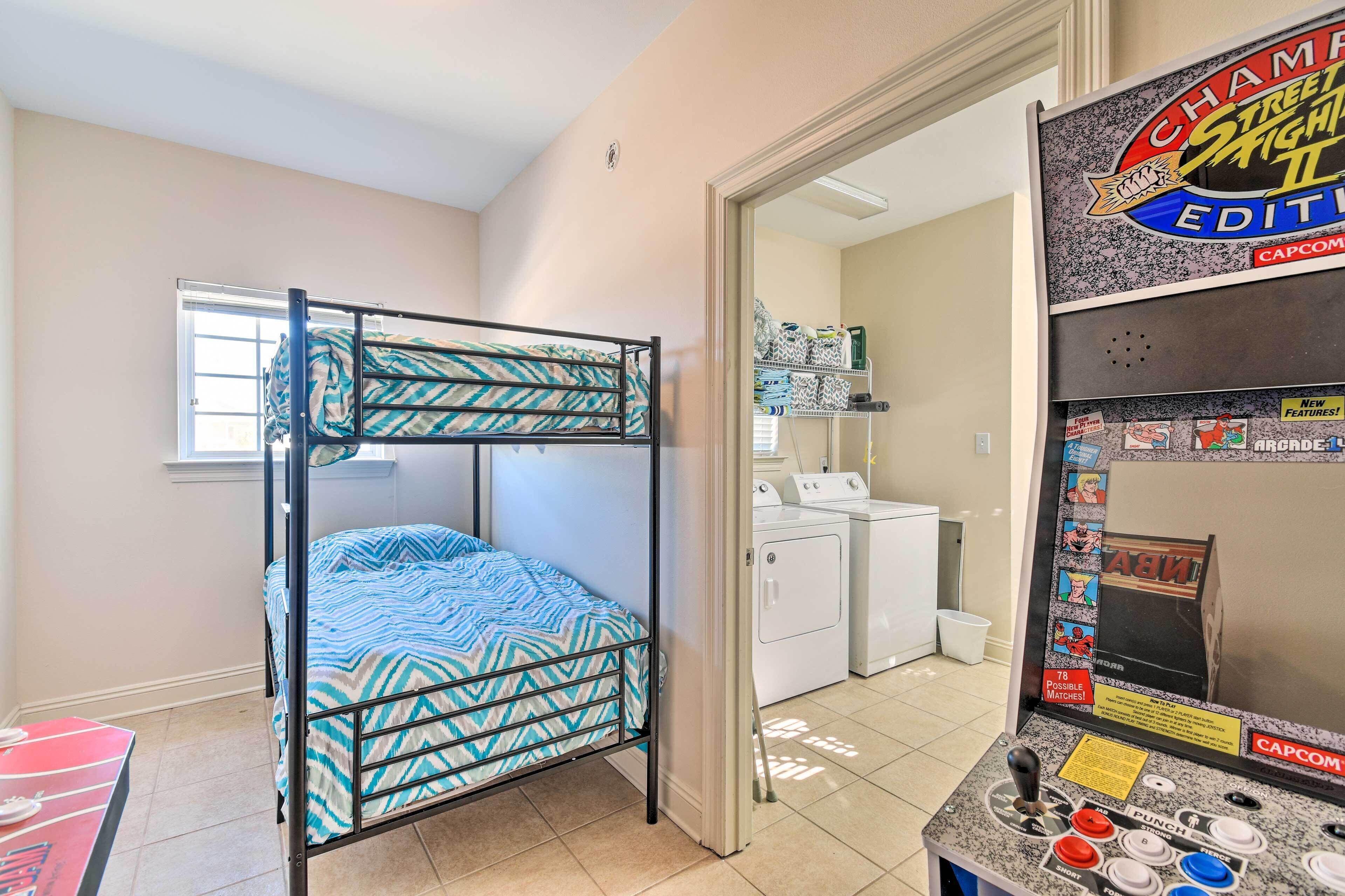 Un lit superposé avec une couvre-lit à motifs, un lave-linge et une borne d’arcade Capcom dans une pièce dotée d’une fenêtre et d’une porte donnant sur une autre pièce.