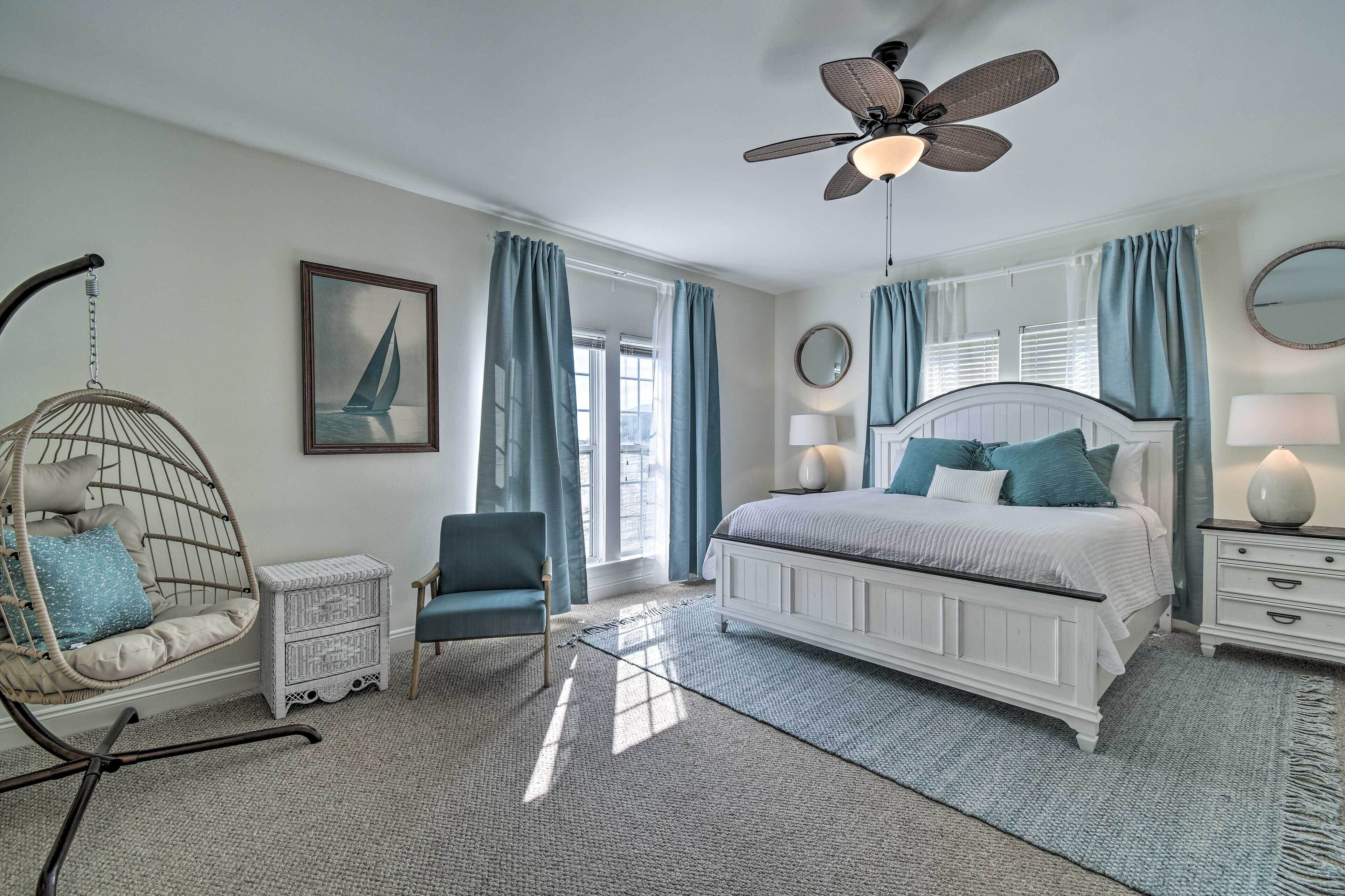 A bedroom with a bed, a hanging chair, a chair, a nightstand, a lamp, a ceiling fan, and a window with curtains.