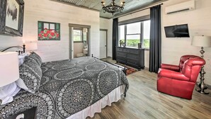 Quarto com cama, poltrona de couro vermelha, lustre, televisão e janela com cortinas.