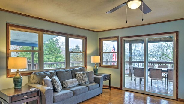 A living room with a gray sofa, two lamps, a wooden coffee table, and a view of an outdoor deck through large windows. - Table Rock Lake House w/ Fire Pit: Steps to Water! (Branson West)
