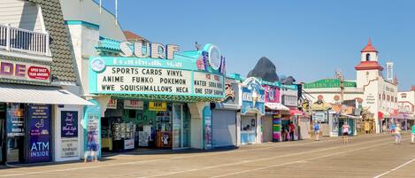 A boardwalk with various shops, including a surf shop, and a historic building with a red roof.