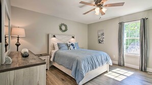 Une chambre à coucher comprenant un lit, une table de chevet, une commode, un ventilateur de plafond, une fenêtre avec des rideaux et un luminaire fixé au mur.