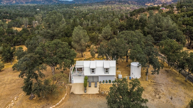 A single-story house with a flat roof, surrounded by dense trees and hills in the background. - Mariposa Home: Furnished Patio & Sierra Mtn Views (Mariposa)