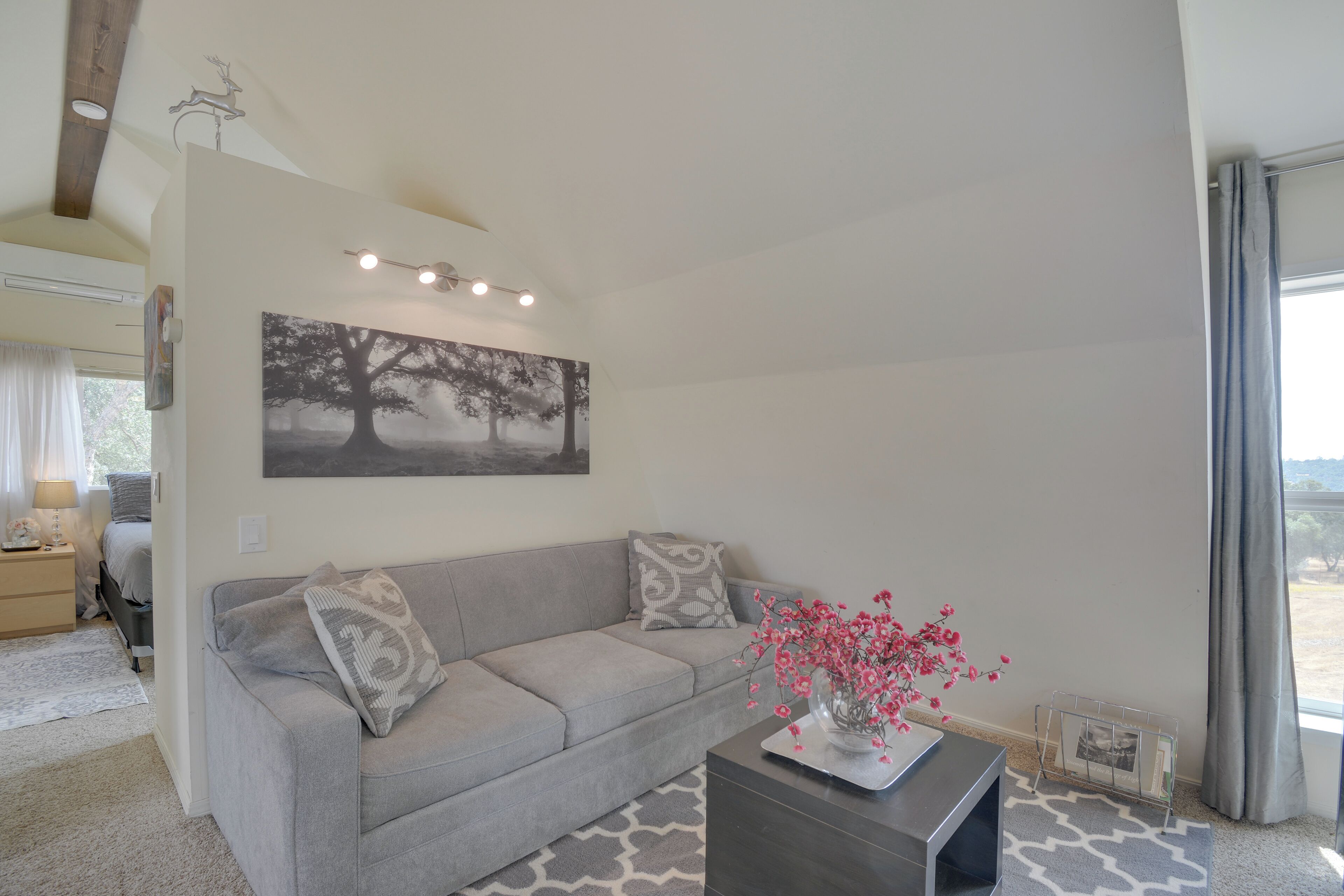 A modern living room with a grey sofa, a coffee table with a vase of flowers, a wall-mounted picture, and a window with curtains.