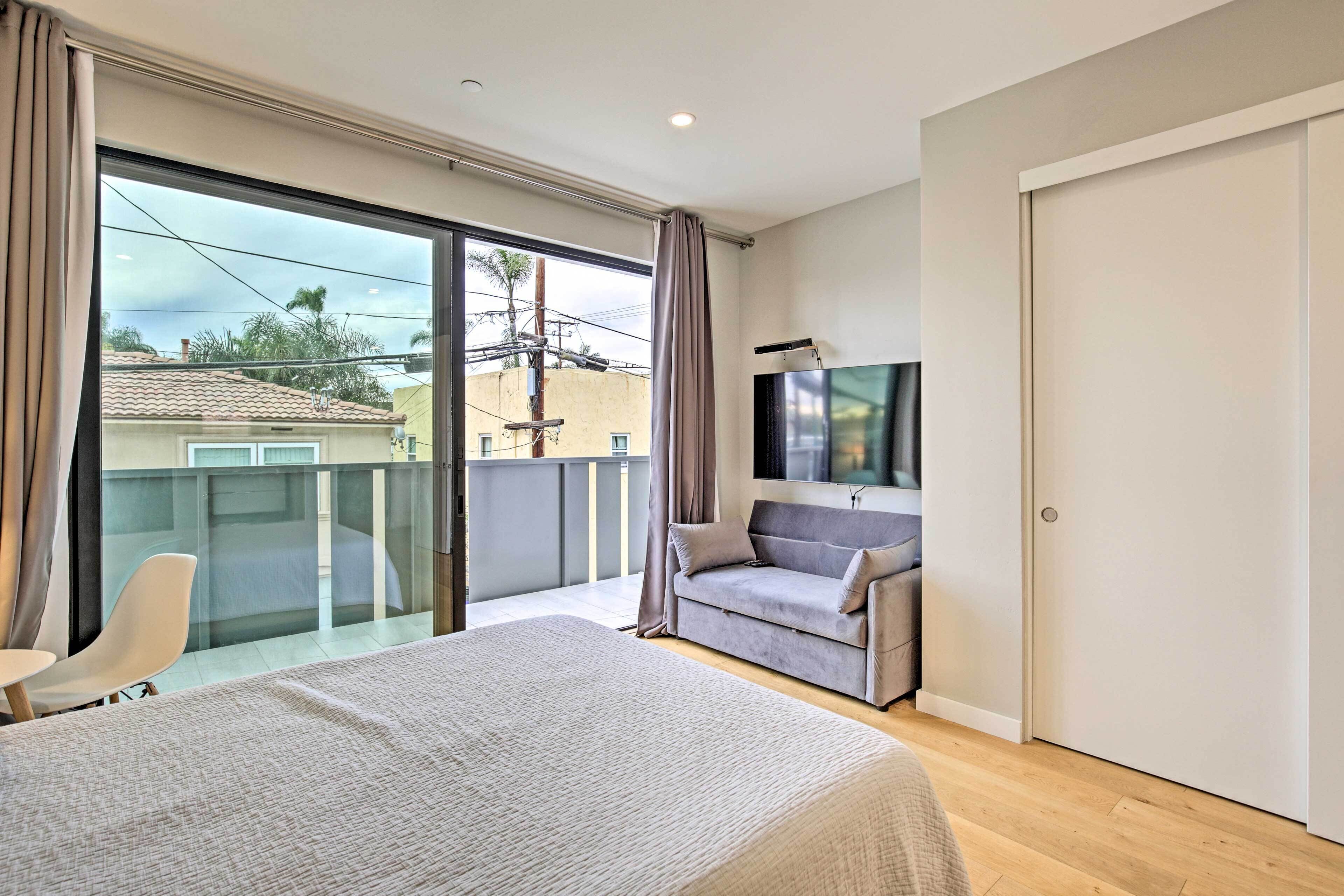 A bedroom with a large bed, a grey sofa, a TV, and a balcony with a view of buildings and power lines.
