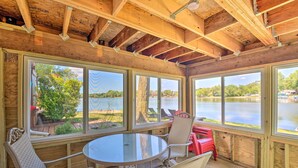 A covered outdoor area with a round table and four chairs, large windows offering views of a lake and trees, and a wooden ceiling structure.