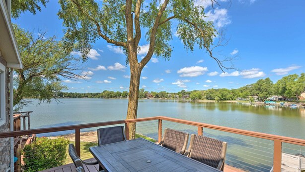 A deck with a table and chairs overlooking a lake, surrounded by trees and a clear sky.