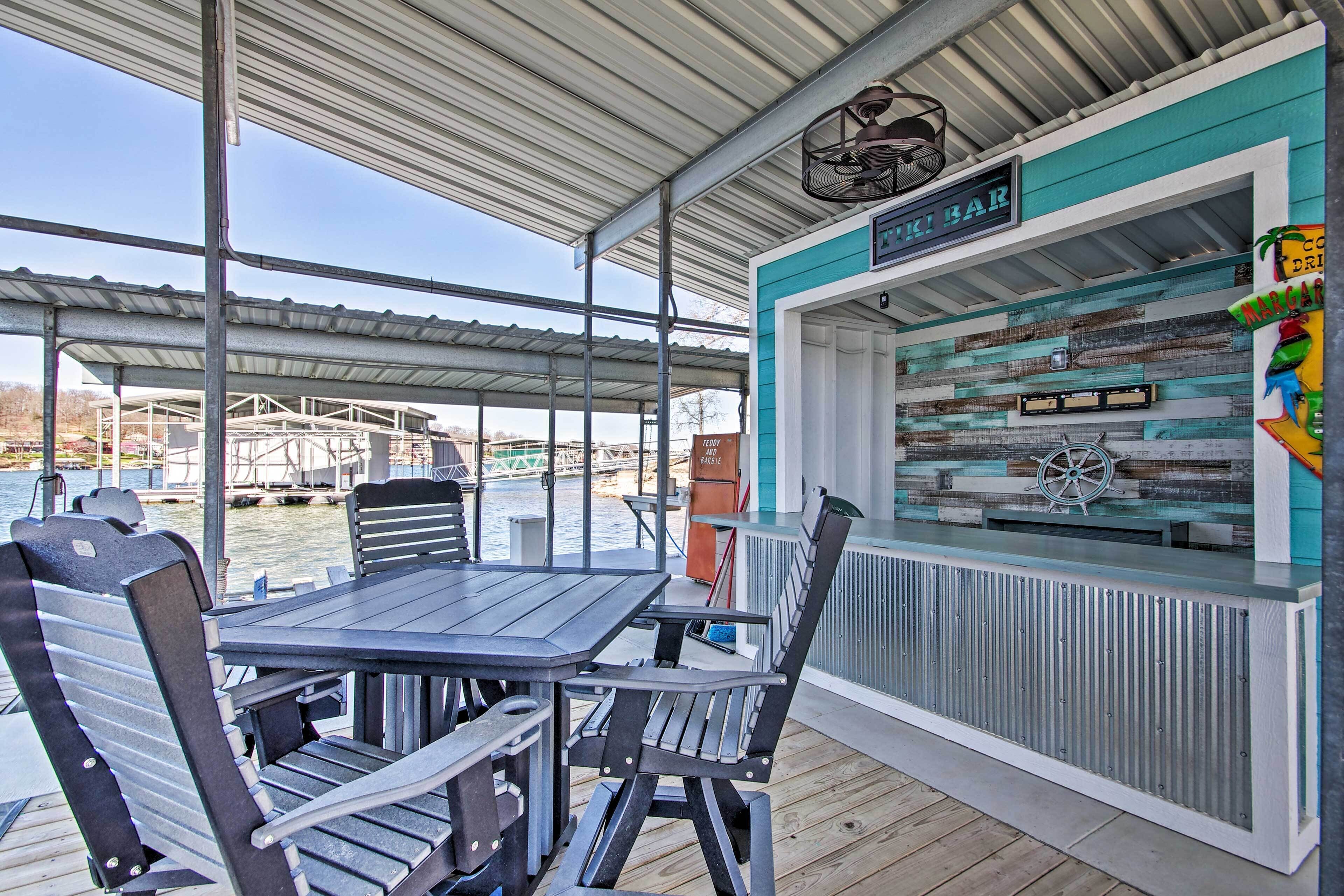 A covered outdoor seating area with a table and chairs, a bar named "The Bar", and a view of the water in the background.