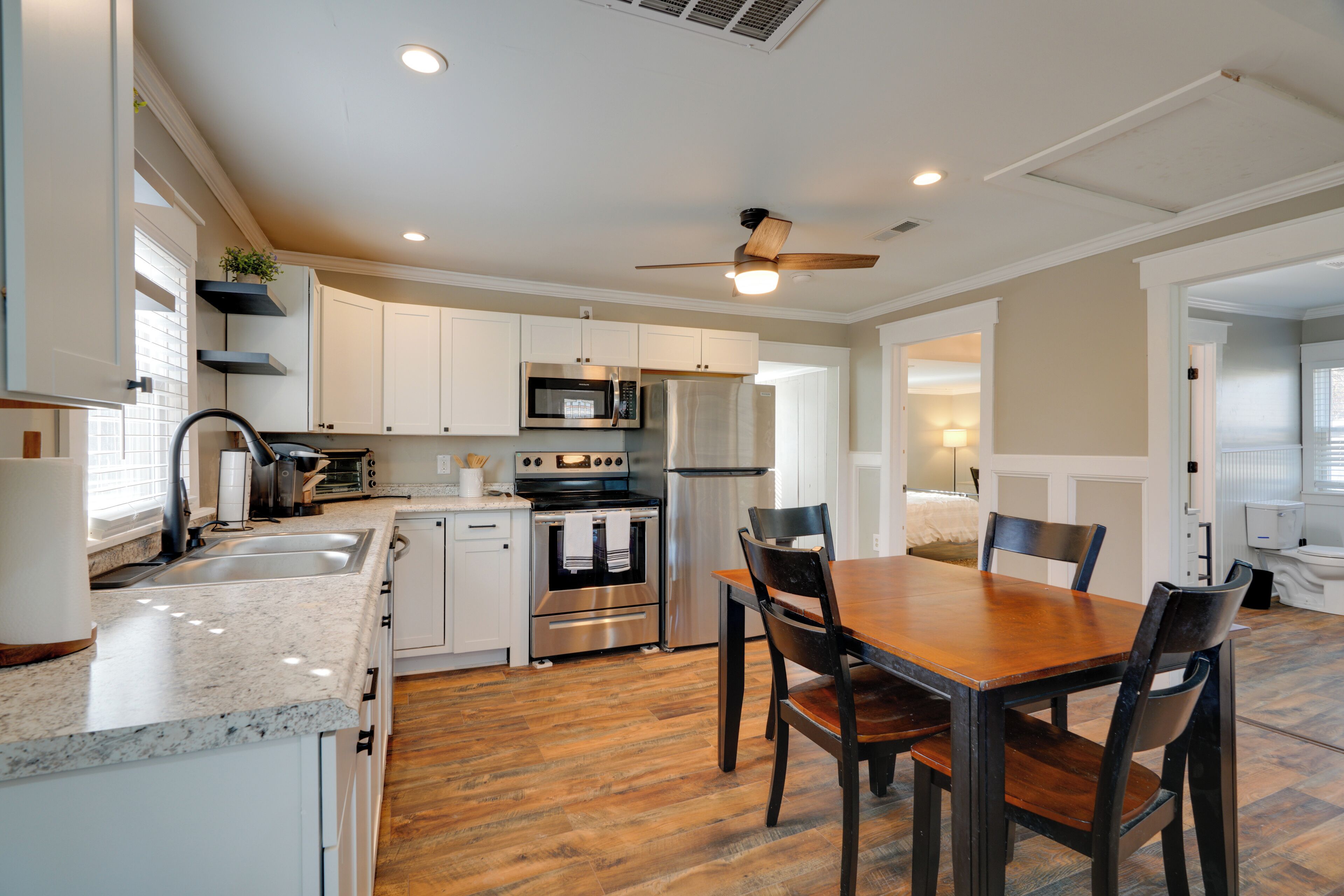 A modern kitchen with white cabinets, stainless steel appliances, a wooden dining table, and a ceiling fan.
